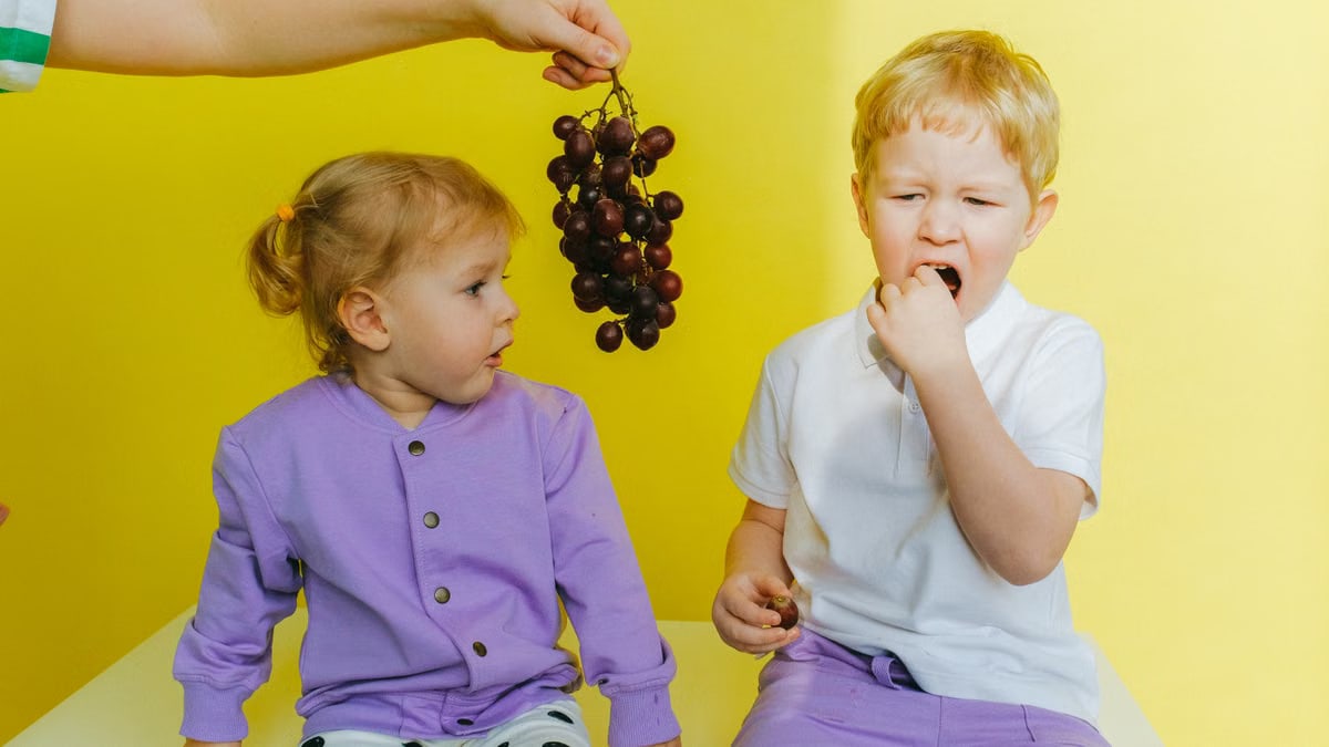 A boy enjoying grapes while sitting next to his curious sister indoors against a yellow background.